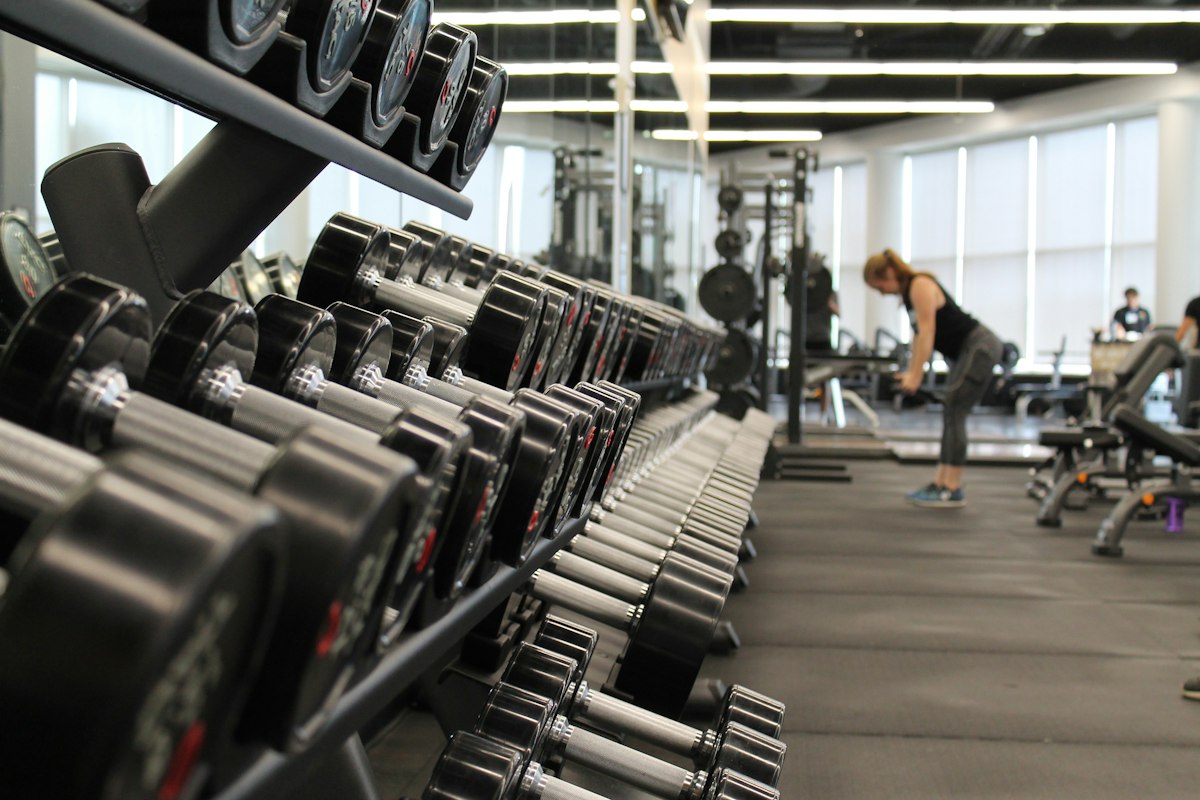 Cinematic photo of a modern strength training gym floor with a bench press station loaded with weight plates, dumbbell rack, kettlebells, battle ropes and a treadmill, dramatic warm overhead lighting, polished mirror wall, empty and spacious, no people